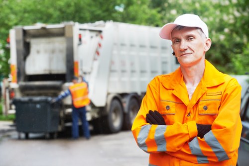 A man wearing a high-visibility orange jacket with reflective stripes and a white cap stands with his arms crossed in the foreground, smiling slightly. Behind him is a large, silver waste collection truck with its rear compartment open, revealing the interior used for rubbish collection. The truck is parked on a paved driveway or street, with a second worker in a high-visibility vest near the rear loading area, placing or removing black rubbish bins. The environment is outdoors with green foliage and trees visible in the background, suggesting a residential or suburban area. The scene appears to be part of a rubbish removal service, with the focus on waste collection activity typical of Hampstead House Clearance services operating in the surrounding London postcode area near NW3 or NW5. The image conveys a professional waste management operation with municipal or private rubbish clearance in progress.
