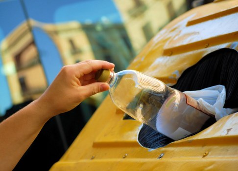 A close-up view of a person's hand using a screwdriver to remove a plastic bottle from a yellow vehicle, likely a waste collection or rubbish removal vehicle, with part of the vehicle's surface visible in the foreground. In the background, blurred buildings suggest an urban environment, possibly in Hampstead or surrounding areas in North West London. The scene captures the process of clearing rubbish, with the plastic bottle partially inserted into a designated opening on the vehicle's body, which appears to be made of metal with a smooth, glossy finish. The lighting indicates natural daylight, enhancing the clarity of the objects and surroundings, emphasizing the focus on waste collection activities typical of Hampstead House Clearance and local rubbish removal services.