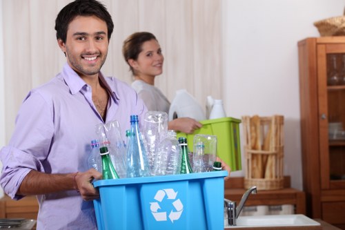 A young man with dark hair and a friendly expression is standing in a well-lit kitchen, holding a large blue recycling bin filled with empty glass bottles of various shapes and sizes, including clear and green-tinted glass. He is wearing a light purple shirt and has his left hand supporting the side of the bin, while his right hand grips the edge. Behind him, a woman with light brown hair tied back is smiling and looking toward the camera, standing near a green plastic storage box on a kitchen counter. The background features wooden cabinets, a wicker basket, and a beige curtain, indicating a domestic environment. The overall scene suggests an environmentally conscious waste collection activity, with a focus on glass recycling, consistent with domestic rubbish removal services provided by Hampstead House Clearance in the local area.
