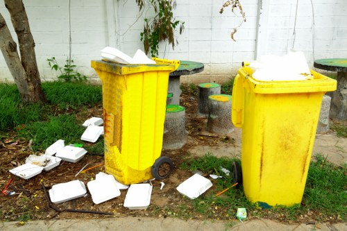 Two large yellow wheelie bins placed on a grassy verge beside a concrete pathway, with scattered white foam food containers and some trash on the ground around them. The bins are made of plastic with textured surfaces, and both have black wheels and lids, one of which is open. In the background, there is a white wooden fence with visible vertical planks, some green foliage and a small tree trunk on the left side. The setting appears to be an outdoor public or residential area typical of urban or suburban Hampstead, with loose litter indicating a recent rubbish collection or waste accumulation. The scene is illuminated by natural daylight, creating a clear and neutral visual context related to rubbish disposal and waste management services that Hampstead House Clearance provides in London or nearby postcode areas.