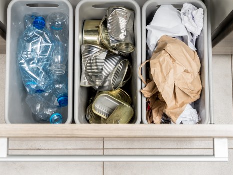 The image displays a three-compartment waste sorting station, likely situated in a kitchen or utility area. The left compartment contains several clear plastic bottles, mostly crushed or flattened, with blue caps visible on some. The middle compartment is filled with various aluminum cans, some crumpled and others neatly stacked, in silver and gold tones. The right compartment holds crumpled paper bags and pieces of white paper, some loosely folded and others slightly crumpled, all surrounded by a neutral-toned container. The containers appear to be made of plastic with smooth surfaces and are arranged side by side, fitting within a light-coloured countertop or work surface, possibly in a domestic or commercial setting in Hampstead or nearby. The environment includes a tiled floor with light, neutral-coloured tiles beneath the station, and the area is well-lit with even lighting, emphasizing the contents designated for recycling or rubbish collection. Hampstead House Clearance’s waste management services are relevant here for helping with the proper disposal or collection of household rubbish in the local area.