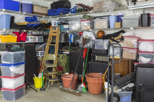 A cluttered storage area filled with various household rubbish and clutter, including multiple transparent plastic storage bins stacked on metal shelving, some with red and black lids, containing miscellaneous items. In front of the shelving, there are two large terracotta plant pots, a metal hand truck, and a wooden step ladder leaning against the shelves. A yellow bucket and cleaning tools, such as mops and brooms, are also visible among the clutter. The area appears to be a garage or storage room, with a concrete floor and fluorescent lighting, consistent with a typical household waste collection or clearance scenario, such as those handled by Hampstead House Clearance for domestic rubbish removal services in the Hampstead area.